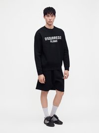 Νέος άνδρας φοράει μαύρο φούτερ DSQUARED2 Milano, μαύρα σορτς, λευκές κάλτσες και μαύρα αθλητικά παπούτσια Adidas, στέκεται μπροστά σε λευκό φόντο.