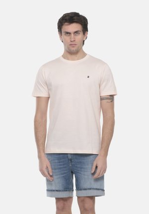 Uomo che indossa una t-shirt a maniche corte rosa chiaro e pantaloncini di jeans blu risvoltati, in piedi davanti a uno sfondo bianco.