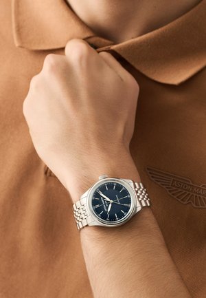 Muñeca con reloj de metal plateado que marca las 9:14, descansando sobre el pecho y vistiendo una camiseta polo marrón con el logo bordado de Aston Martin.