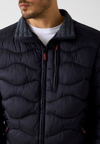 Marineblaue Steppjacke mit Steppmuster und hohem Kragen. Mit roten Reißverschluss-Zippern und kontrastierendem grauem Kragenfutter. Zwei Seitentaschen.