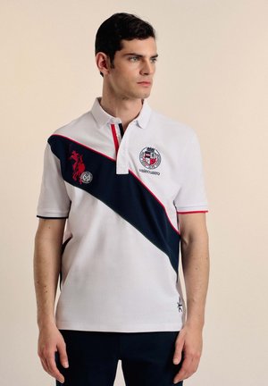Camisa polo blanca con un panel diagonal en azul marino, detalles en rojo y logotipos bordados. Mangas cortas con ribete de contraste en los puños.