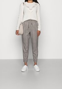 Pull blanc légèrement texturé avec bordure en dentelle, associé à un pantalon gris à carreaux et des baskets blanches. Le mannequin tient un petit sac beige.