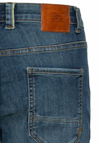 Jeans en denim bleu foncé avec des coutures jaunes. Présente une étiquette en cuir beige portant la marque "camel active" sur la ceinture arrière. Une poche arrière.