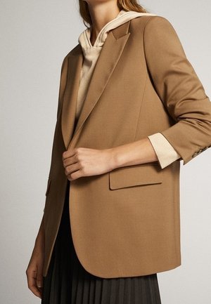 Kobieta ubrana w karmelową marynarkę na beżowej bluzie z kapturem, z czarną plisowaną spódnicą, widoczna od szyi do połowy uda na jednolitym tle.