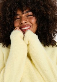 Joven con cabello rizado que sonríe cálidamente, vestido con un suéter suave de color amarillo pálido, con las manos parcialmente cubriendo su rostro en un gesto acogedor.