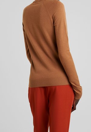 Person iført en tætsiddende lysebrun langærmet sweater og løse røde bukser, set forfra mod en ensfarvet grå baggrund.