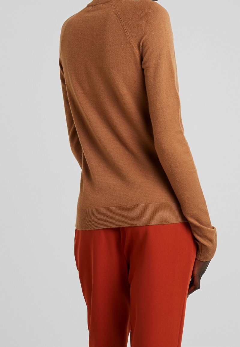 Personne portant un pull ajusté marron clair à manches longues et un pantalon rouge ample, vue de dos sur un fond gris uni.