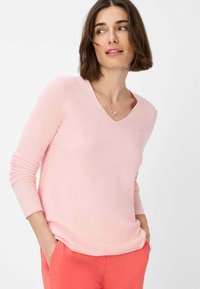 Helles rosa Langarmshirt mit V-Ausschnitt und geripptem Muster. Das Kleidungsstück ist locker geschnitten, mit Händen in den Taschen, und wird mit korallenfarbenen Hosen kombiniert.