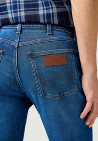 Jeans de mezclilla azules con una etiqueta de cuero marrón en el bolsillo trasero, que presentan un distintivo patrón de costura en zigzag y detalles de remaches estándar.