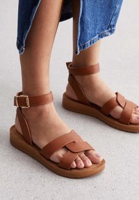 Des sandales en cuir marron avec des sangles tressées, une sangle à boucle à la cheville et une semelle en bois légère. Design à bout rond ; texture lisse.