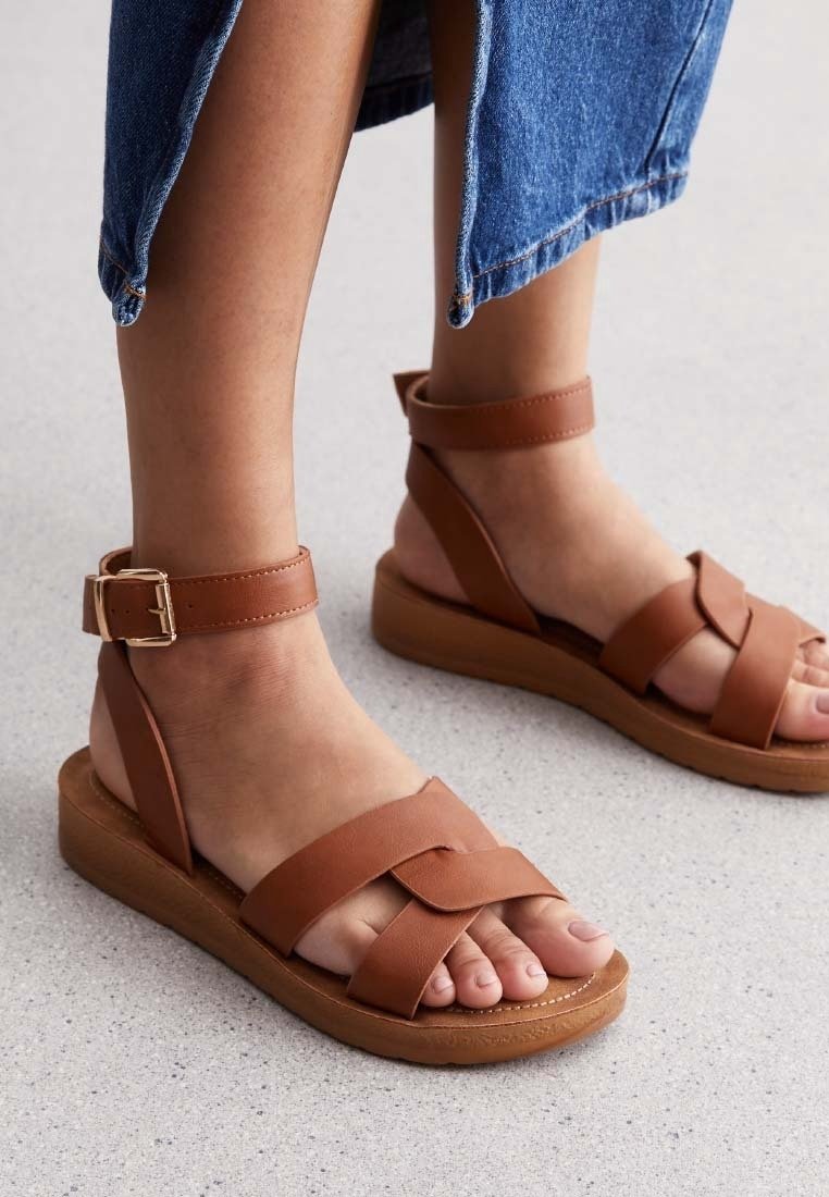 Des sandales en cuir marron avec des sangles tressées, une sangle à boucle à la cheville et une semelle en bois légère. Design à bout rond ; texture lisse.