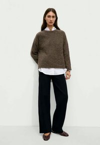 Rjav puhast pulover preko belega srajce, v kombinaciji s širokimi črnimi hlačami in maroon čevlji s konicami. Preprost kroj in sproščen fit.