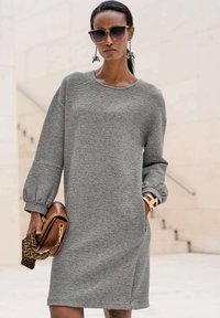 Graues Strickkleid mit langen, puffärmeln und seitlichen Taschen. Glatte Textur. Mit einer braunen Handtasche und goldenen Armreifen accessorized.