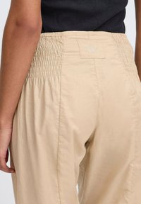 Beige nadrág, smocked derékkal, amelynek finom fényessége és könnyű textúrája van. A hátsó oldalon látható a "PULZ JEANS" címke.