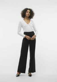 Top bianco a maniche lunghe con scollatura a V e texture arricciata, abbinato a pantaloni neri a gamba larga su misura e décolleté neri a punta.