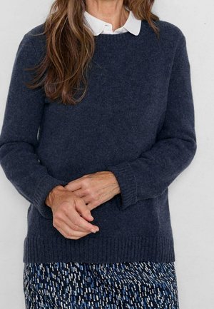 Kvinde iført marineblå sweater over hvid skjorte med krave og mønstrede bukser, står med hænderne løst foldet foran sig.