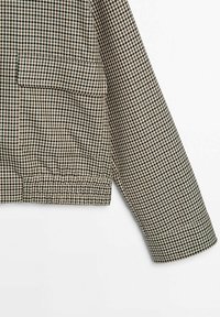 Manche et bas de veste à carreaux beige et noir avec ourlet élastique et poche rabat avant sur fond uni.