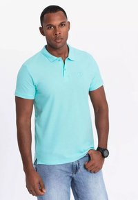 Camisa polo de color azul claro hecha de un tejido texturizado, con cuello, mangas cortas y un logo sutil en el pecho. Combinada con unos jeans azules.