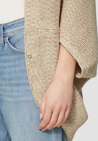 Hellbeige gestrickte Strickjacke mit einem strukturierten Muster und weiten Ärmeln, kombiniert mit hellblauen Jeans. Detail umfasst Knopfbeschläge.