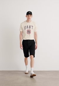 Cream t-shirt with "GANT" logo, black shorts, white socks, and brown sneakers with black accents. Simple, casual outfit displayed.