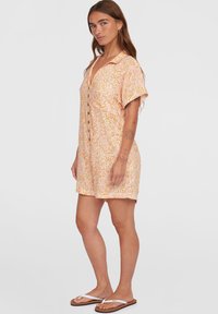 Robe à manches courtes et boutonnée en pêche clair avec un motif floral ; possède un col et une coupe décontractée. Portée avec des tongs marron.