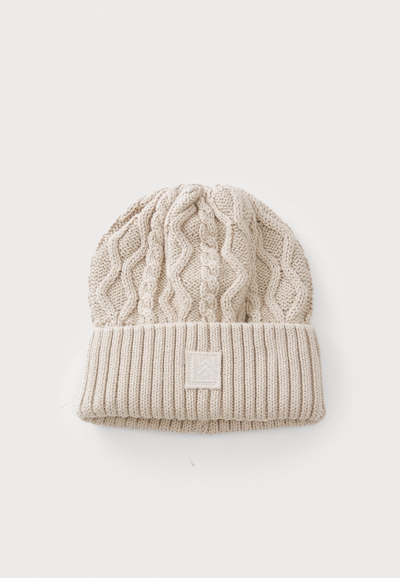 Beige strikket lue med kabelstruktur på toppen og ribbet mansjett. Har et lite firkantet logo-patch foran. Mykt, koselig materiale.