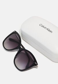 Ochelari de soare negri cu rame supradimensionate și unghiulare, și lentile întunecate cu un gradient, însoțiți de un etui alb Calvin Klein cu logo negru.