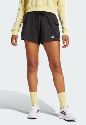 Persoon die zwarte Adidas short met zakken draagt, gele sokken, witte Adidas sneakers en een geel sweatshirt met witte strepen op de mouwen.