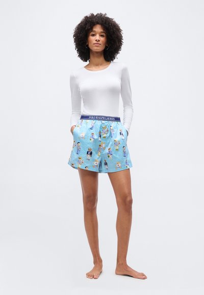 Γυναίκα με σγουρά μαλλιά που φορά λευκό μακρυμάνικο τοπ και μπλε ριγωτή βερμούδα με σχέδια αρκουδάκια και ζώνη Polo Ralph Lauren.