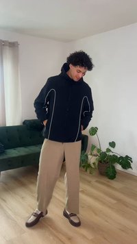 Νέος άνδρας με μαύρο μπουφάν με λευκή επένδυση, μπεζ παντελόνι και καφέ μοκασίνια, όρθιος μέσα σε εσωτερικό χώρο σε ξύλινο πάτωμα, κοντά σε πράσινο καναπέ και φυτά σε γλάστρες.