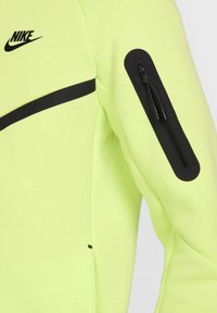 Chaqueta Nike con un tejido amarillo-verde brillante, detalles en negro y un bolsillo con cremallera en el brazo. Textura suave y diseño atlético.