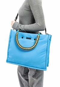 Blauwe gequiltte draagtas met een gevlochten handvat, voorzien van oranje en turquoise details, een rechthoekige vorm en een gladde, lichte textuur.