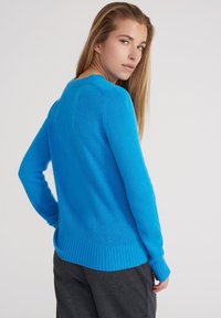 Hellesblaues Strickpullover mit runder Halslinie, gerippten Bündchen und Saum, der eine weiche Textur und einen entspannten Schnitt aufweist, kombiniert mit dunkelgrauen Hosen.