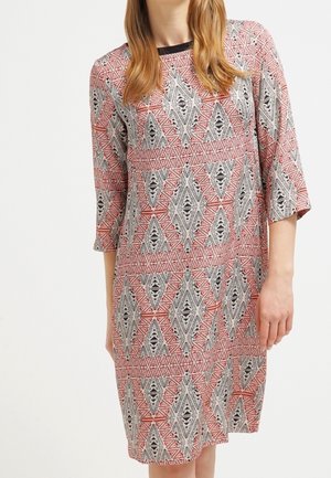 Kobieta ubrana w sukienkę do kolan z długim rękawem, ozdobioną geometrycznym wzorem diamentów w kolorach czerwonym, czarnym i białym oraz z czarną lamówką na kołnierzu.