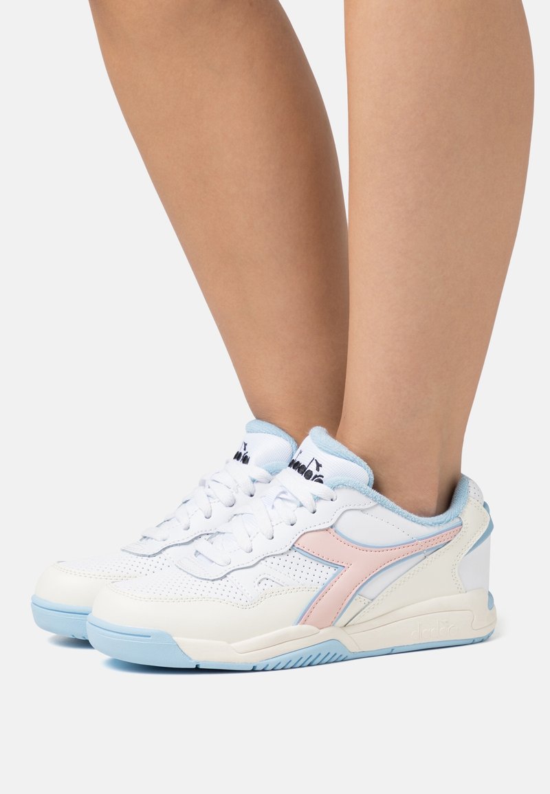Des jambes portant des baskets blanches avec des semelles bleu clair et des logos latéraux roses, debout sur un fond blanc uni.