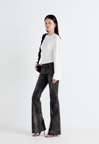 Model stands confidently in a fitted white long-sleeve top and flared dark pants, with a neutral background enhancing the outfit.