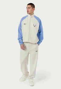 Hellgraue Trainingsjacke mit blauen Ärmeln und weißen Hosen. Enthält einen Frontreißverschluss, Logo-Stickerei sowie elastische Bündchen und einen elastischen Bund.