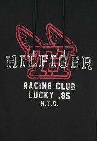 Čierna mikina s kapucňou s veľkou červenou grafikou s krídlami a slovom "HILFIGER." Text obsahuje "RACING CLUB LUCKY .85 N.Y.C." v bielej farbe.