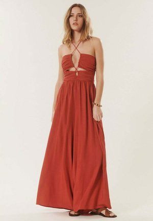 Femme portant une robe maxi sans manches rouge rouille avec des découpes et des bretelles croisées, assortie à des sandales marron et un bracelet.