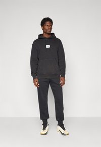 Sort hættebluse med kængurulomme og grøn logo. Kombineret med mørke sweatpants og højhælede sneakers med cremefarvede og sorte detaljer.