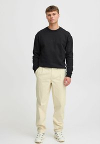 Zwarte sweatshirt met getextureerd logo op de borst, gecombineerd met crèmekleurige, op maat gemaakte broek. Model draagt witte sneakers met een subtiel patroon.