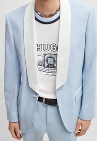 Veste de costume bleu clair avec un revers en satin blanc, fermeture à un bouton, associée à un t-shirt graphique blanc et une ceinture noire. Tissu texturé.