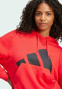 Röd hoodie i mjukt material med en stor svart grafisk tryck och en känguruficka, med dragsko på huvan och avslappnad passform.