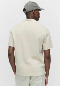 Lyhythihainen, kermaisen värinen polo paita, jossa on tekstuuri. Ominaisuuksiin kuuluu kaulus ja rento istuvuus, ja se on yhdistetty vaaleanvärisiin housuihin sekä harmaaseen lippalakkiin.