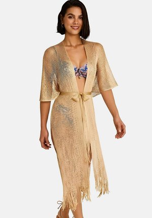 Femme portant un couvre-chef beige à ceinture en maille ouverte avec des franges sur un haut de bikini à fleurs, souriante et debout devant un fond blanc.