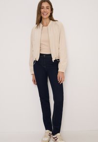 Beige bomberjack met elastische manchetten, gecombineerd met donkerblauwe recht-uitlopende jeans. Crème sneakers met rode en marineblauwe accenten maken de outfit compleet.