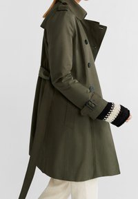Olivengrüner Trenchcoat mit einem doppelreihigen Design, großen Knöpfen, Schulterklappen und einem taillierten Gürtel. Langer Ärmel wird präsentiert.
