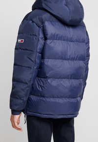 Granatowa pikowana kurtka puchowa z kapturem, z przeszywanym wzorem, elastycznymi mankietami oraz małą naszywką z logo na lewym rękawie. Gładka tekstura.