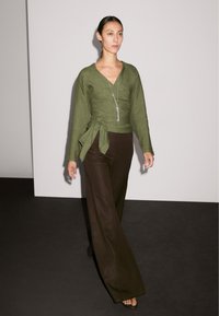 Un mannequin défile avec confiance dans un haut enveloppant vert et un pantalon large marron, mettant en valeur un collier en argent sur un fond minimaliste.