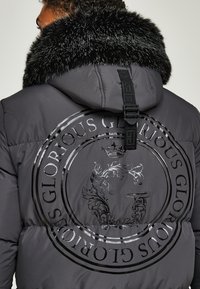 Daunenjacke in anthrazitgrau mit einem Kragen aus schwarzem Kunstfell. Sie verfügt über ein großes, kreisförmiges Grafiklogo auf der Rückseite in glänzendem Druck.